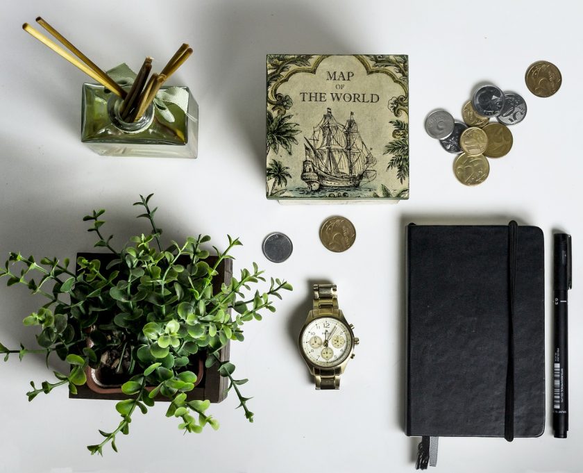 flatlay of white tabletop with plant, watch, bullet journal, pen, coins, small book with words "map of world", and incense in bottle