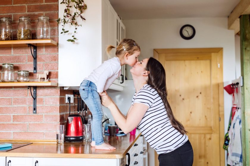 little girl standing on kitchen countertop kissing young mother on the nose