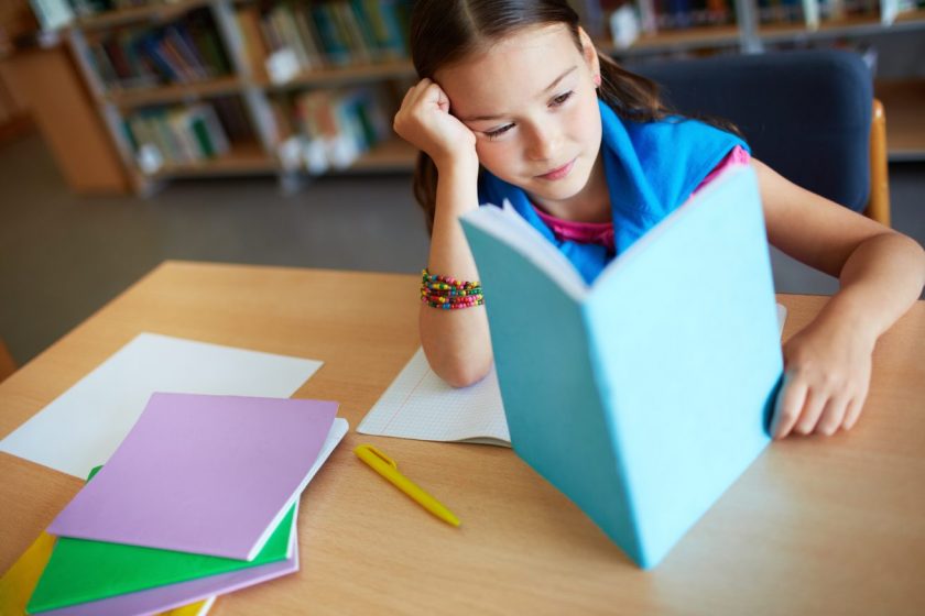 elementary aged girl reading book at desk in library