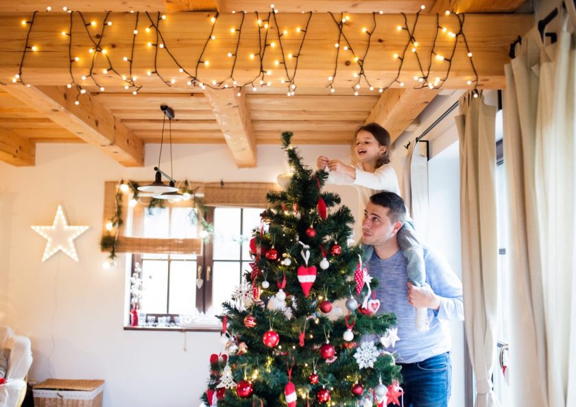 young daughter on dad's shoulders putting ornament on top of the decorated Christmas tree