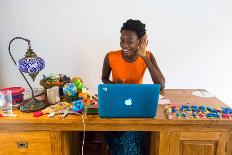 work at home mom VIP teacher sitting at desk facing a computer, smiling with hand to ear