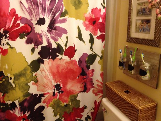 bathroom with bright flowered shower curtain, basket on back of toilet and mason jar toothbrush holder
