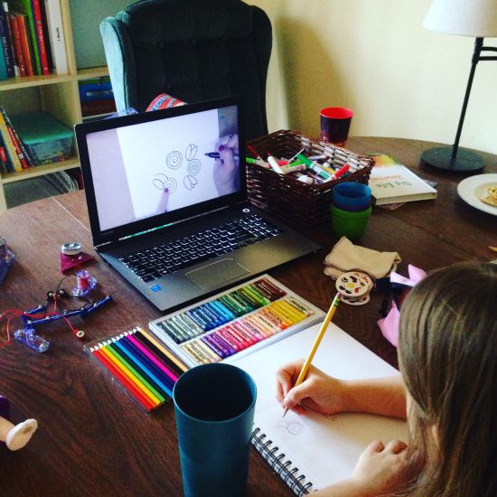 girl sitting at kitchen table with sketch pad, pastels, and colored pencils, watching art tutorial on computer