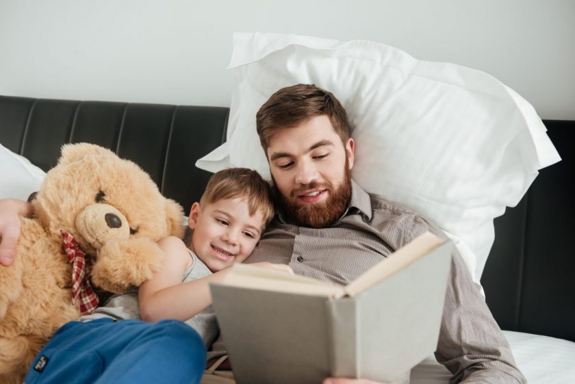 father lying in bed reading picture book to son with giant teddy bear by his side