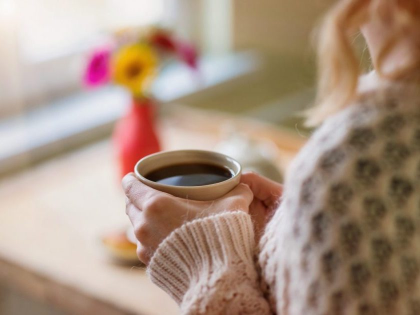 woman in sweater holding cup of coffee, vase of flowers out of focus on table, discouraged about decluttering on low income