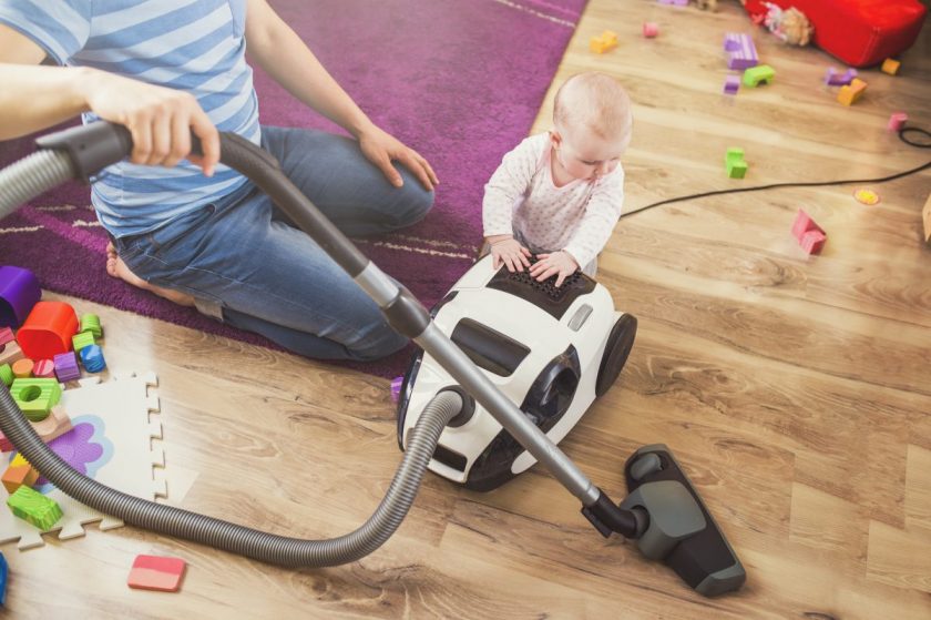 dad trying to vacuum living room with a baby pushing on vacuum, toys scattered everywhere