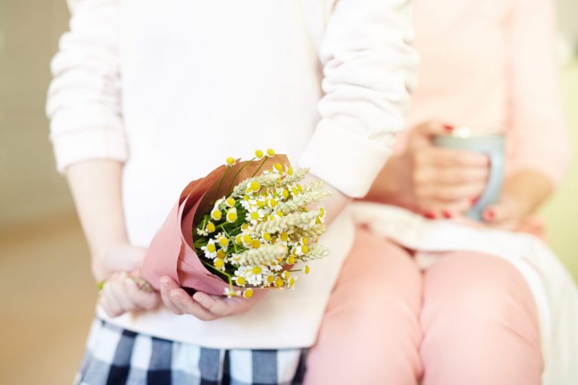 mom sitting with coffee mug with little boy holding flowers behind his back for Mother's Day present