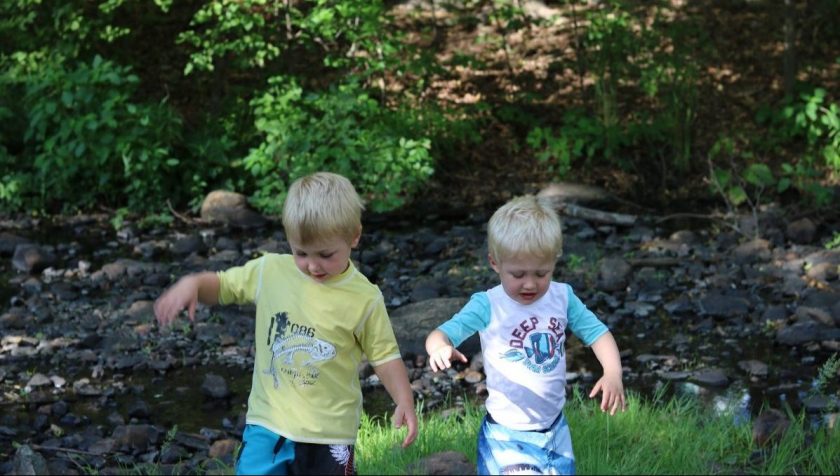 two brothers in swimsuits dipping toes in creek, trees in background