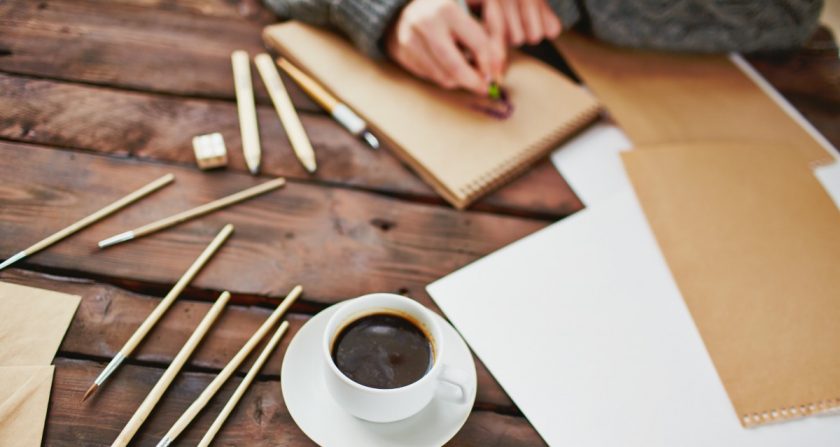 woman writing on book on desk, with coffee, pens and paper all around