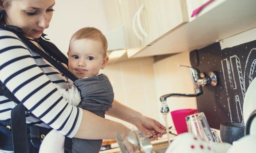 young mom with baby in baby carrier standing at kitchen sink watching dishes