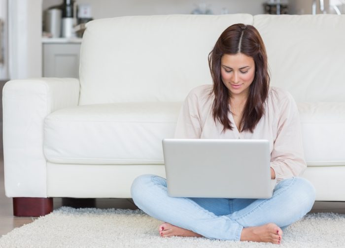 young mom sitting on white rug resting back against white couch with legs crossed and laptop on her lap