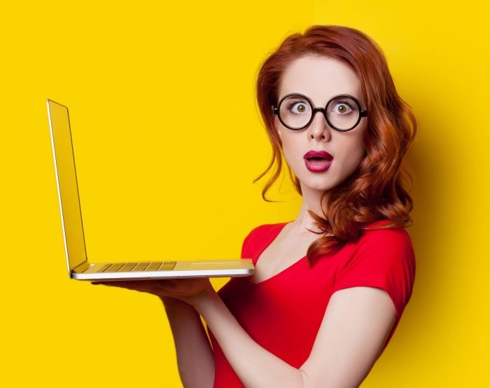 red-headed woman with black glasses holding laptop up in both hands, looking shocked, on yellow background