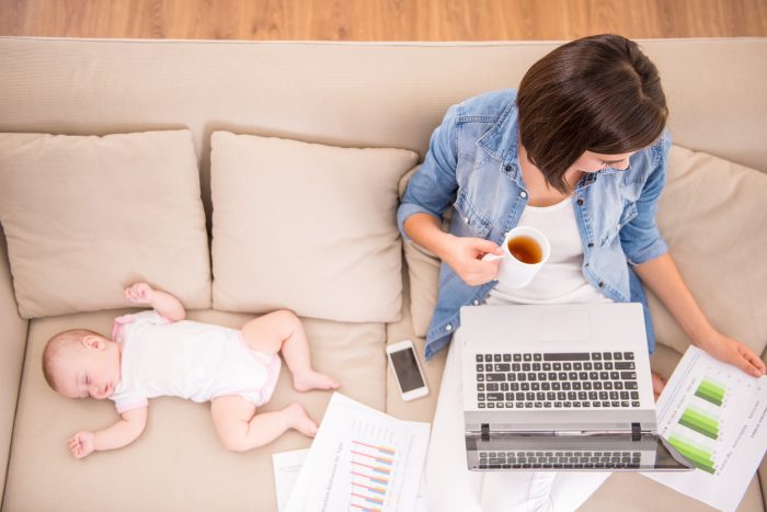 mom blogging with cup of tea in hand, sitting on couch while baby sleeps next to her