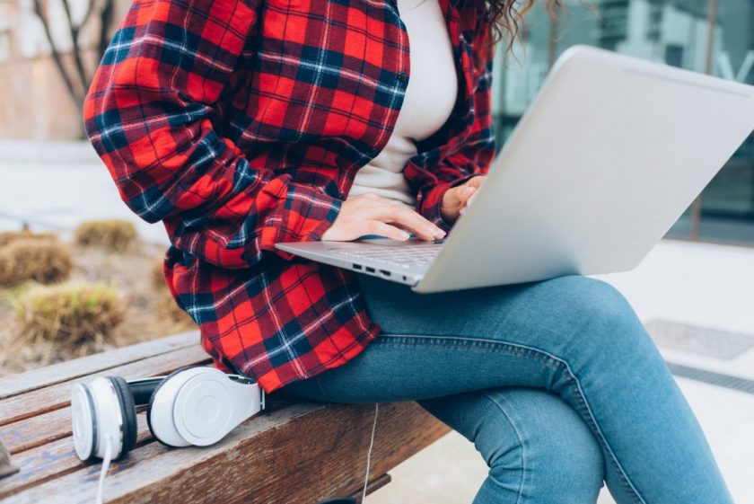 woman in red plaid flannel shirt sitting on park bench with laptop and headphones blogging