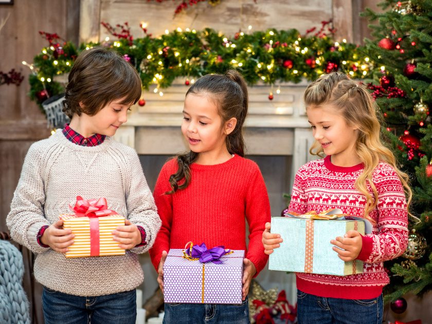 three children in sweaters holding Christmas presents in front of decorated mantle