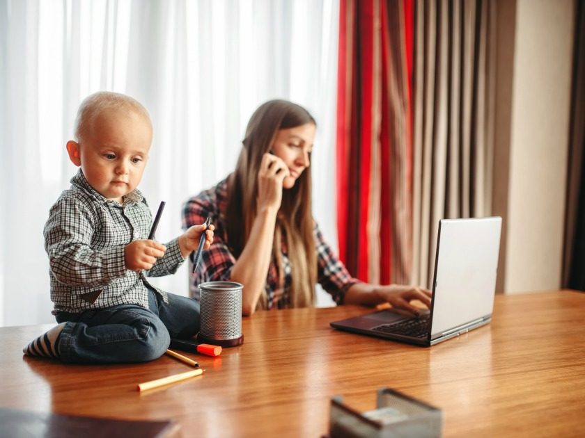 work at home mom typing on computer and talking on phone while toddler sits on top of the table next to her