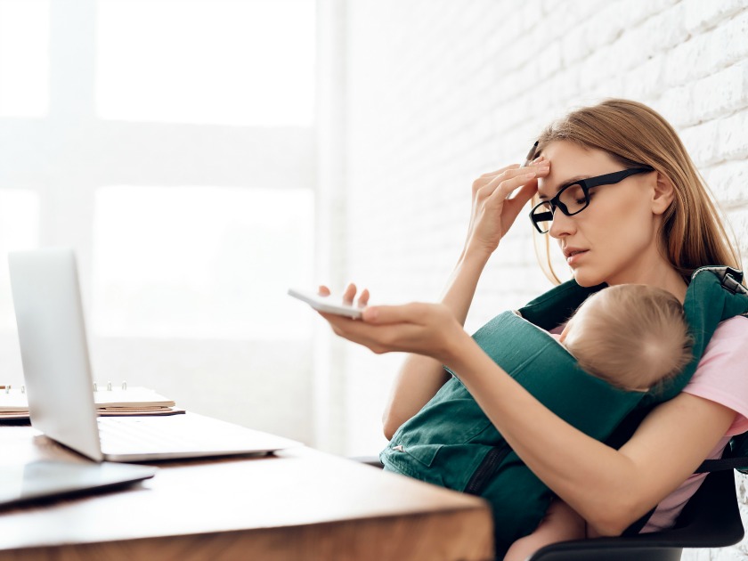 working mom holding head to forehead while sitting at desk with computer, baby asleep in baby carrier on her chest