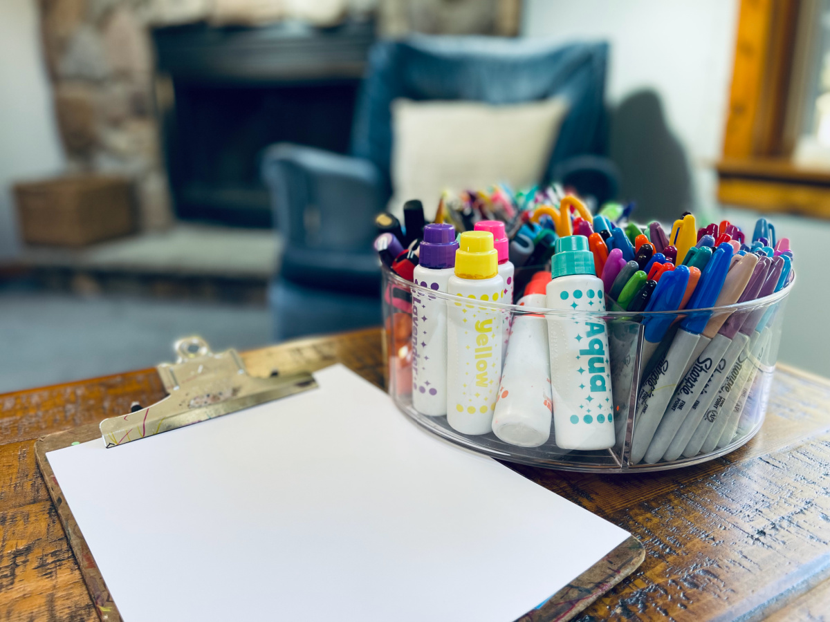 homeschool art supplies in clear acrylic lazy susan on coffee table near clipboard with white paper.