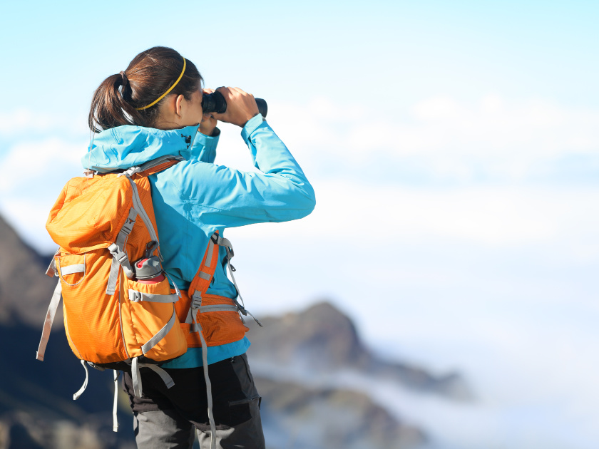 female hiker wearing blue hiking jacket and orange backpack, looking through binoculars on mountain