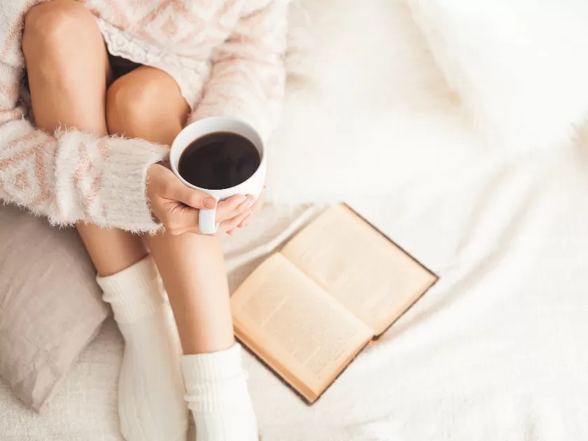 woman in cozy white sweater and socks holding cup of coffee and reading a book on white bedspread