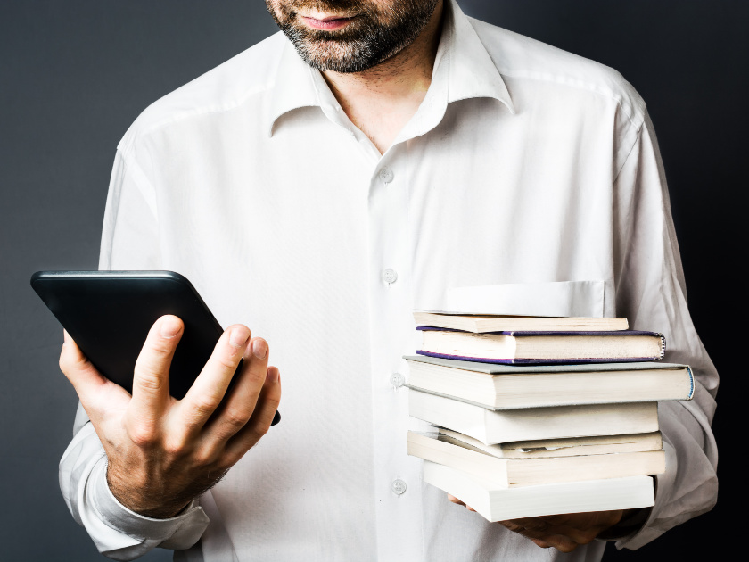 man in button-down collared white shirt looking at kindle in one hand and holding a stack of books in the other
