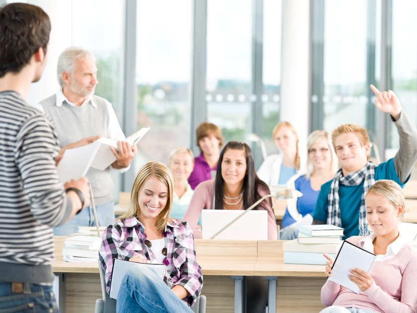 college students in class, taking notes, raising hands and typing on computers