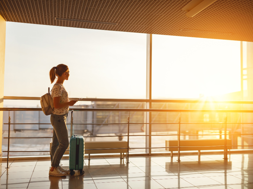 young woman with blue suitcase in airport looking out the window at the sunrise