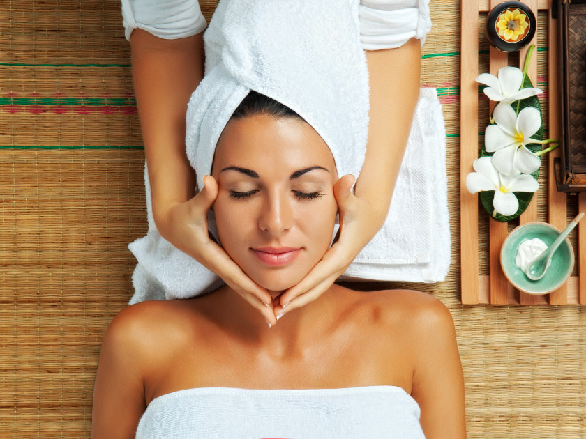 woman with towels on head and body getting facial massage, spa themed tray to her right
