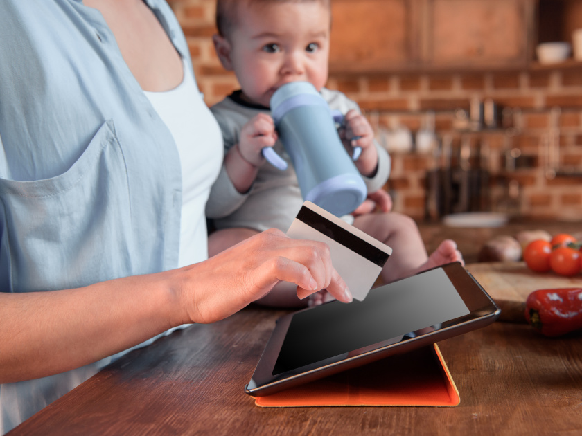 mom with postpartum depression holding baby in one hand and a credit card and tablet in the other