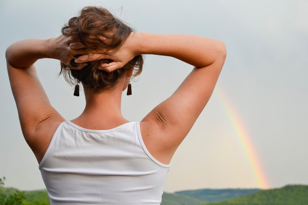 woman looking at rainbow in the distance with hands up behind her head