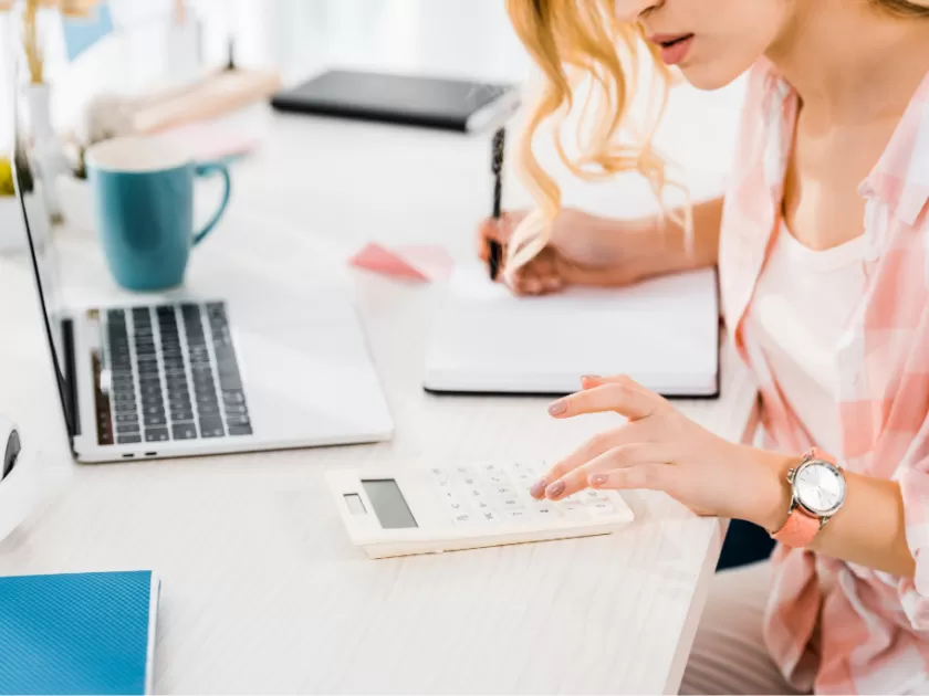 woman at desk writing in notebook and using calculator. laptop in front of her