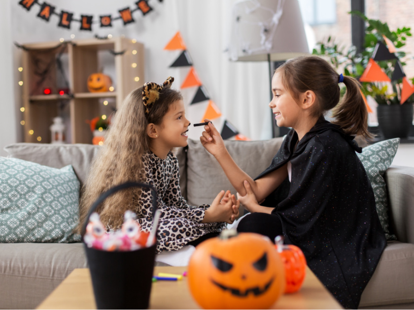 two girls sitting on couch dressing up for halloween, one applying cat makeup to the other