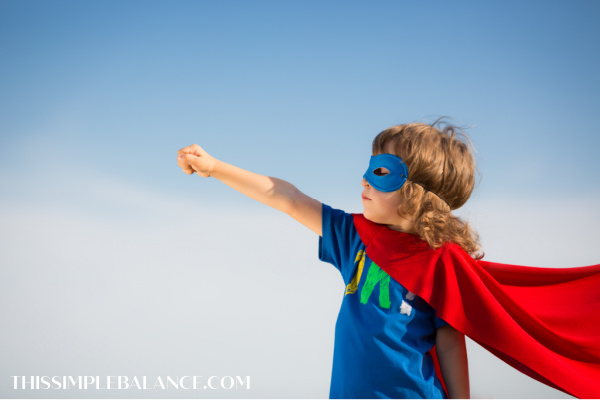 young boy wearing red superhero cape and blue eye mask doing superman stance with blue sky background