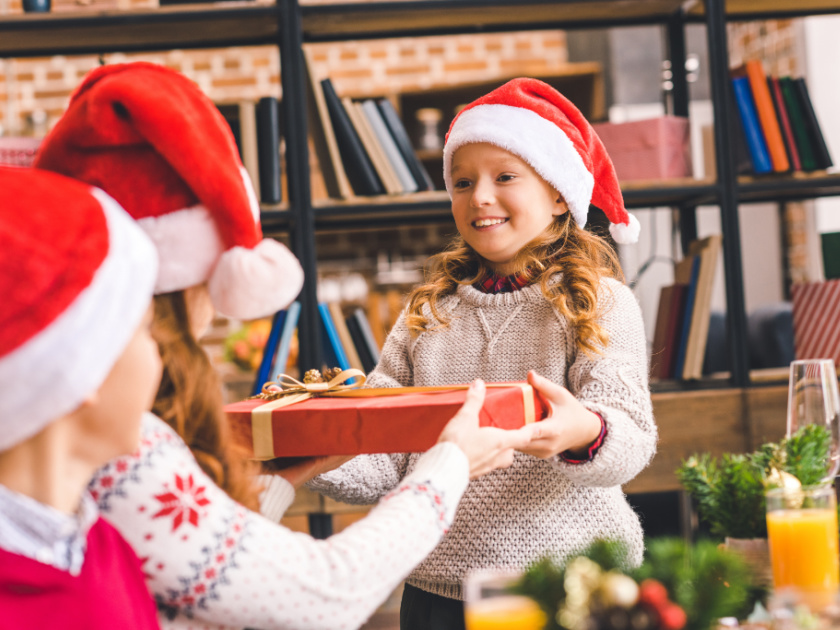 girl with santa hat and white sweater giving wrapped christmas present to mom