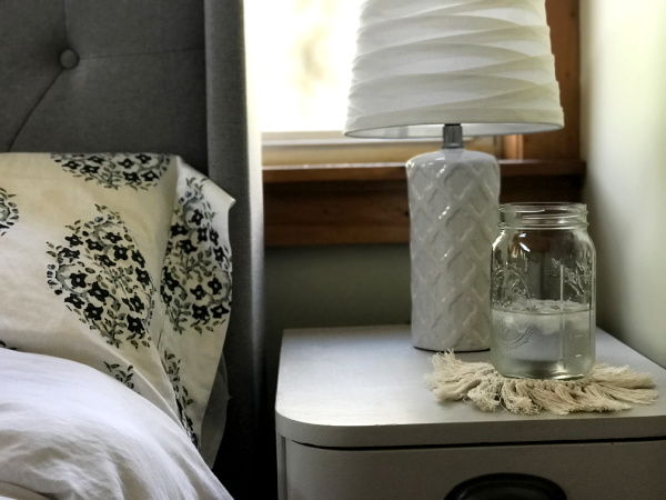 gray bedside table with white lamp, macrame coaster and mason jar with ice water