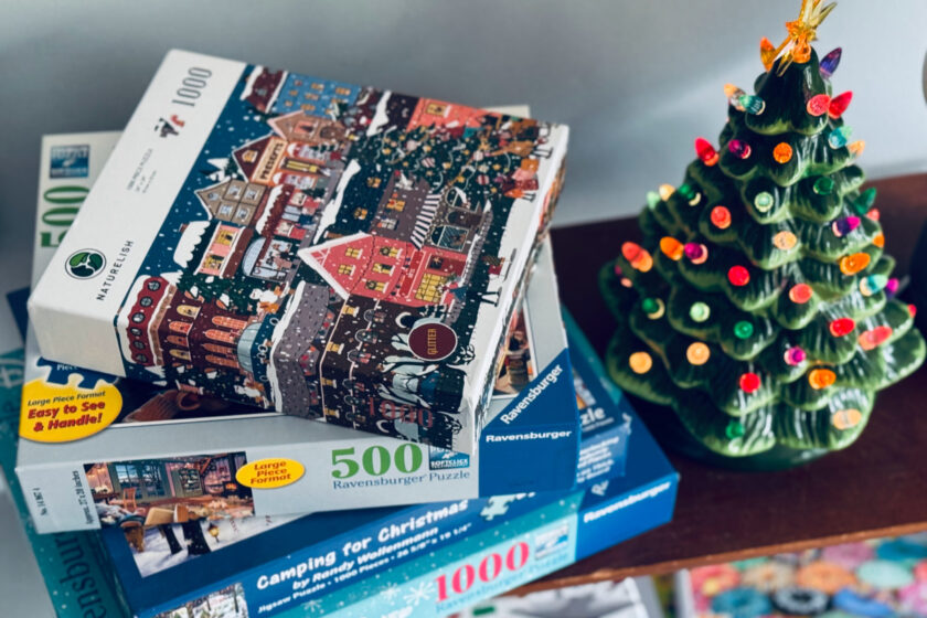 Christmas puzzles stacked on shelf sitting next to ceramic Christmas tree.