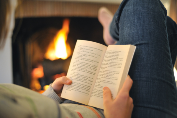 woman laying on couch with feet up reading a book next to a fireplace