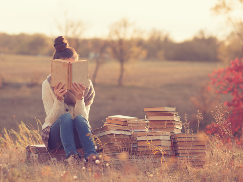 woman sitting on box with her nose in a book about unschooling, and stacks of books beside her, outdoors in meadow