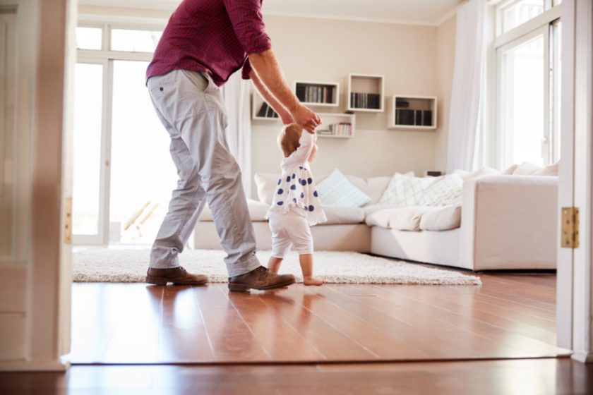 father helping toddler learn to walk in clean white living room