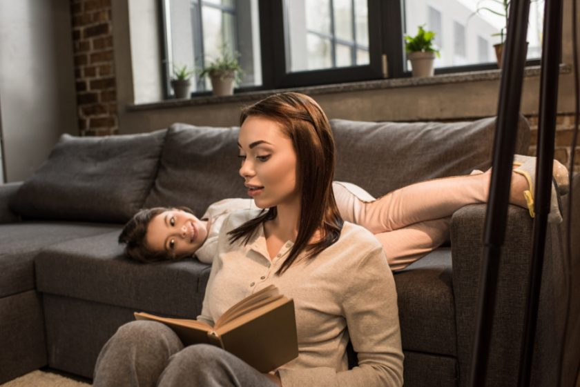 mom sitting on floor reading chapter book to girl lying on couch