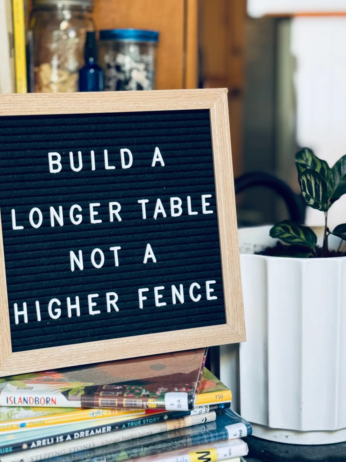 Quote on letterboard near stack of books and plant: "Build a Longer Table, Not a Higher Fence."