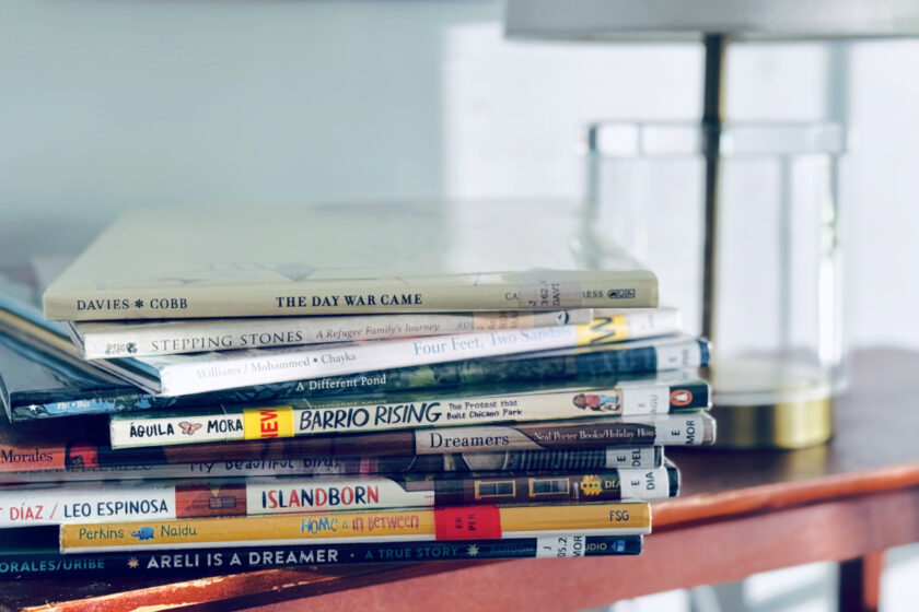 stack of picture books about immigration and refugees on table beside lamp.