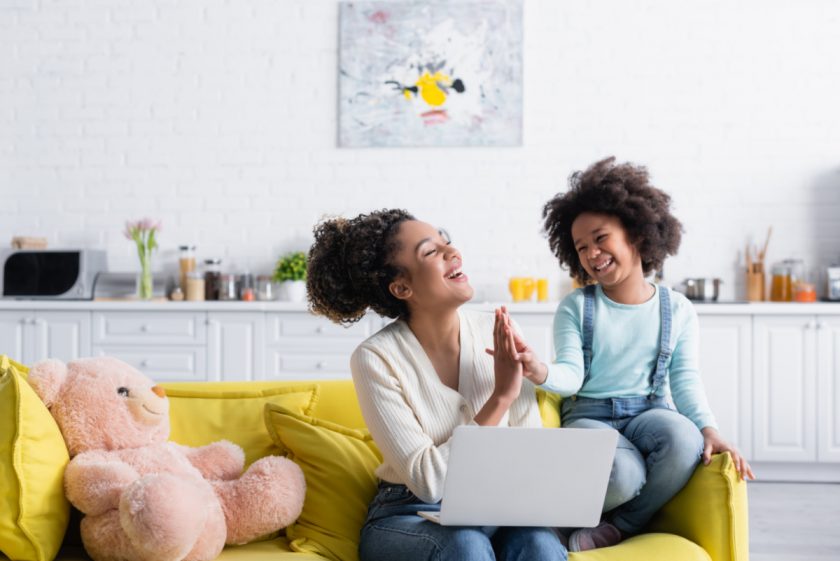 mom sitting on couch working on laptop with child sitting next to her giving a high-five