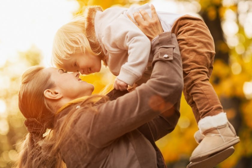 smiling young mom holding blond toddler in the air on a fall day