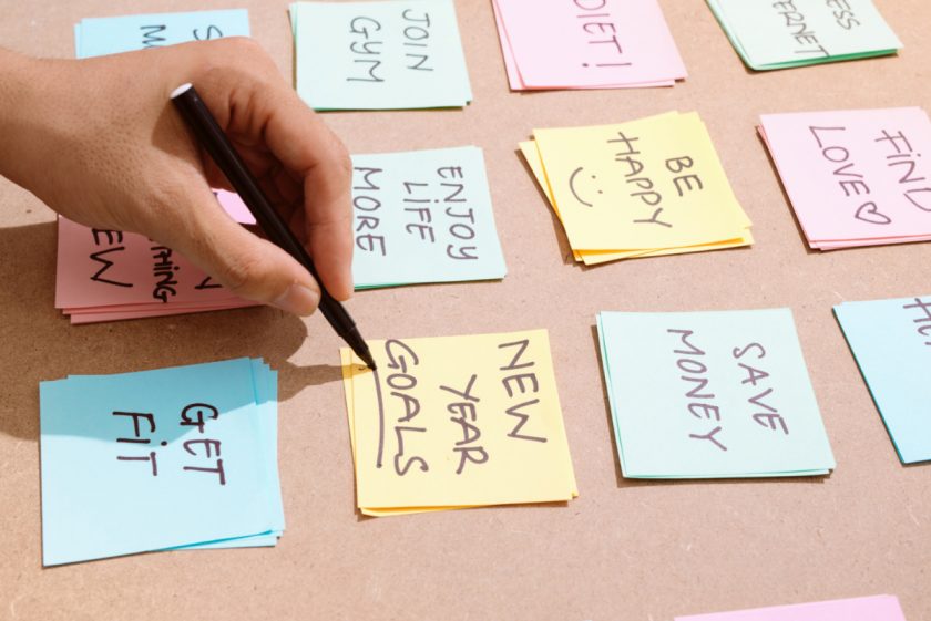 post-it notes surrounded by goals like "save money" and "get fit", with focus on hand underlining the "new years goals" post-it note