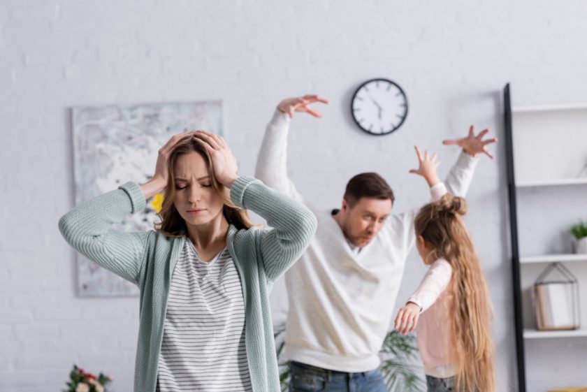 stressed stay at home mom rubbing her temples with eyes closed while husband and daughter play in the background of living room