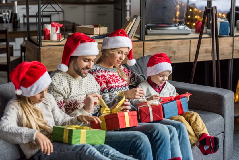family of four wearing santa hats, sitting on gray couch opening christmas presents