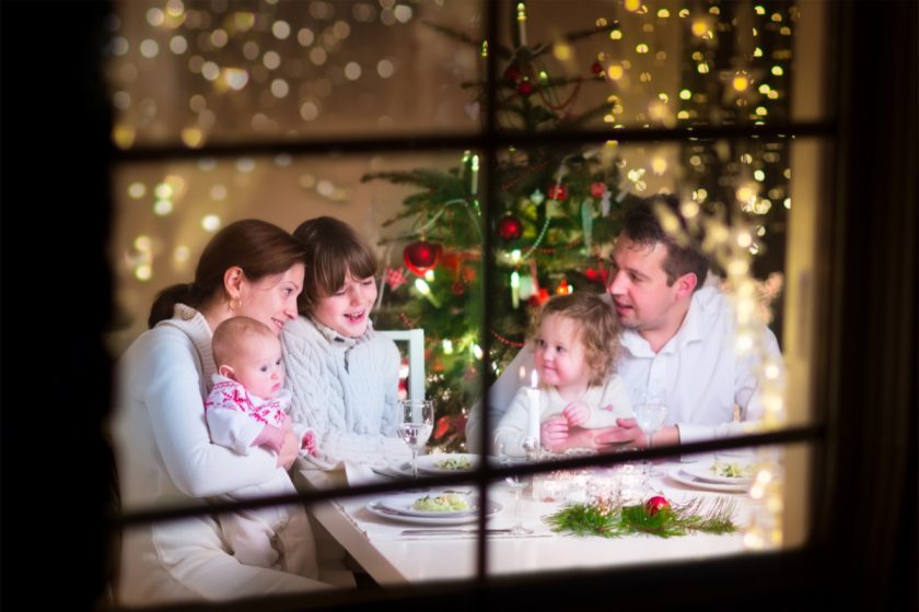 Young big family celebrating Christmas enjoying dinner, view from outside through a window into a decorated living room with tree and candle lights, happy parents eating with three kids