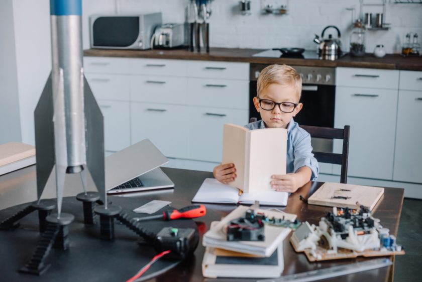 young boy with glasses sitting at kitchen table reading a science book with a model rocket and circuits surrounding him