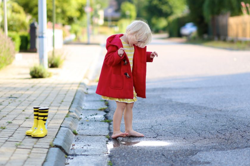 little girl in red coat playing in puddle in bare feet, rain boots on the sidewalk